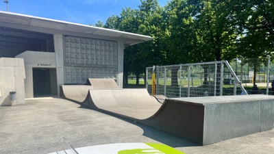 Skatepark mit Halfpipe und grünen Bäumen im Hintergrund.