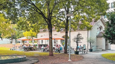 Menschen sitzen in einem Parkcafé unter Bäumen bei sonnigem Wetter.