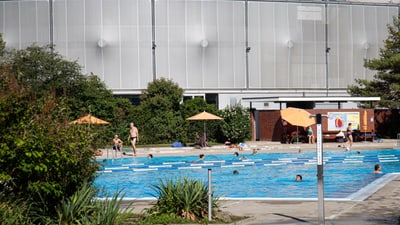 Menschen schwimmen in einem Freibad mit Gebäude im Hintergrund.