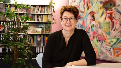 Gaby Hintermannsitzt lächelnd an einem Tisch in einem bunt dekorierten Raum mit Bücherregal und Pflanze.