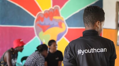 Person mit #youthcan-Shirt vor bunter Wand mit Faustmotiv, andere Personen im Gespräch.