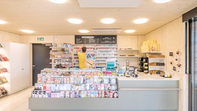 Modern gestalteter Ladeninnenraum mit Regalen voller Zeitschriften und hinter der Kasse steht ein Mann mit gelben T-Shirt.