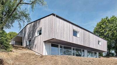 Gebäude mit Holzfassade am oberen Ende eines Hanges mit Bäumen links und rechts.