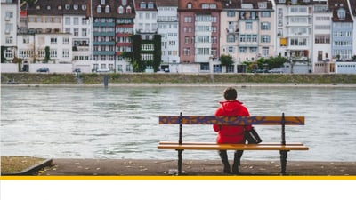 Person in roter Jacke sitzt auf einer Bank am Fluss, mit Gebäuden im Hintergrund.