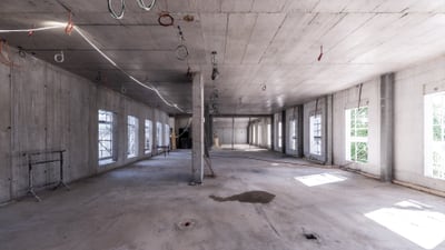 Innenansicht einer rohbauartigen Halle mit Betonwänden und -boden, leere Fensteröffnungen.