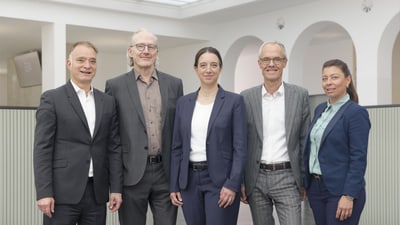Fünf Personen in Businesskleidung stehen nebeneinander in einem modernen Büro.