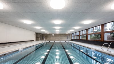 Schwimmbecken mit vier Bahnen. Die Schwimmhalle ist mit weissen Plättli und heller Beleuchtung an der Decke und einer Fensterfront.