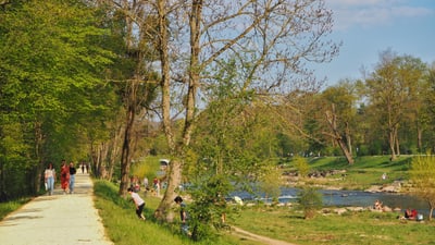 Flussufer mit Weg daneben, viele Leute sitzen im Gras.