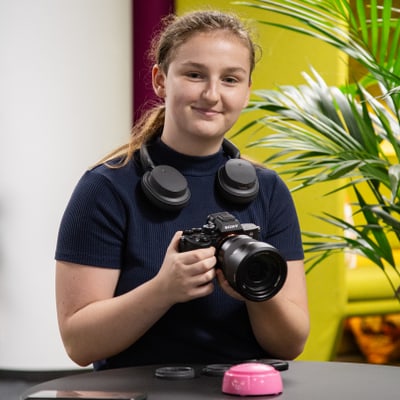 Person mit Kamera und Kopfhörern an einem Tisch sitzend.