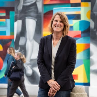 Lotti Lienhard posiert vor bunter Wandmalerei, Menschen im Hintergrund.