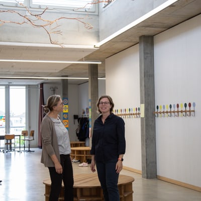 Schulleiterin Patricia Hacker und Projektleiterin Dagmar Schüneman stehen in modernem Raum mit Wandkunst und grossen Fenstern.