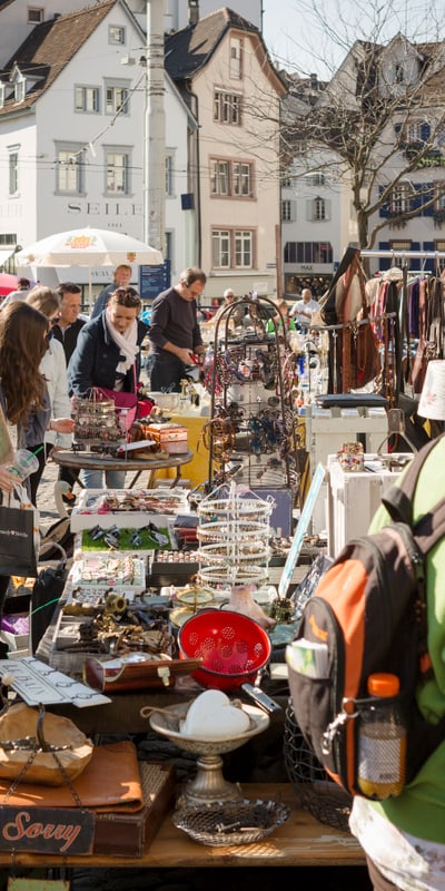 Menschen auf einem Flohmarkt mit verschiedenen Ständen und Gebäuden im Hintergrund.