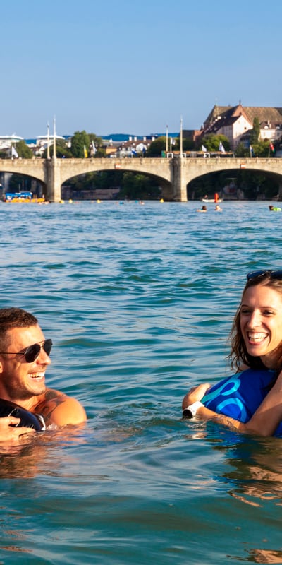 Menschen schwimmen im Fluss mit einer Brücke und Stadt im Hintergrund.