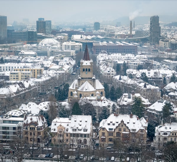 Verschneite Stadtansicht mit Kirche im Vordergrund und Hochhäusern im Hintergrund.