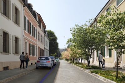 Strasse mit geparktem Auto und Spaziergängern zwischen Wohnhäusern und Bäumen.