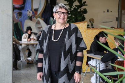 Sandra Bettoni mit Brille steht in einer Kunstschule, Studenten im Hintergrund.