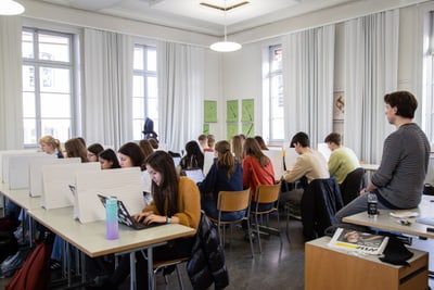 Schüler im Klassenzimmer am Computer, Lehrer sieht zu.