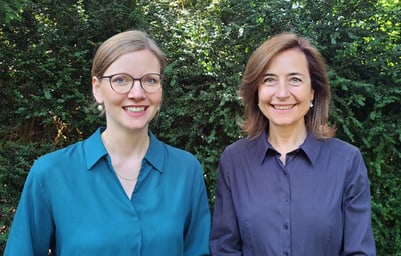 Zwei lächelnde Frauen vor Büschen im Freien.