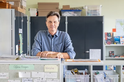 Person in blauem Hemd steht vor einem Schreibtisch mit Regalen und Büromaterial.