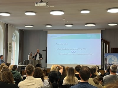 Mann hält Präsentation vor Publikum in einem Raum mit Projektor.
