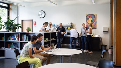 Personen in einem Pausenraum mit Bücherregalen und Tisch beim Kaffee zubereiten und unterhalten.