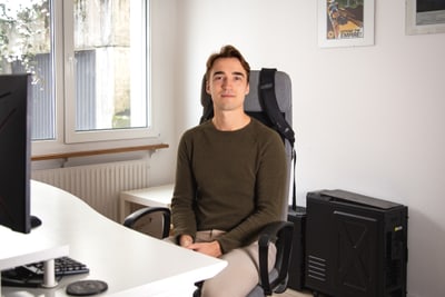 Aljosha Gasser in grünem Pullover sitzt an Schreibtisch in Büro mit Computer.