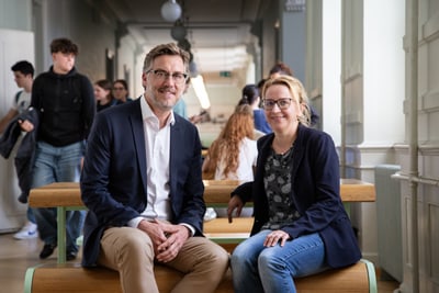Patrick Langloh und Judith Hindermann sitzen auf einer Bank in einem Flur, andere Menschen im Hintergrund.