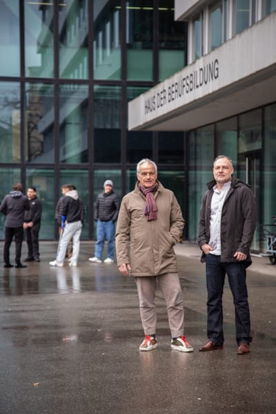 Benedikt Arnold und Matthias Hostettler vor einem modernen Gebäude mit mehreren Personen im Hintergrund.