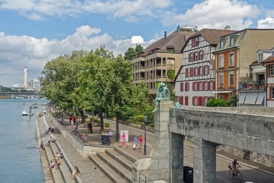 Stadtszene mit Fluss, Bäumen und Menschen entlang der Promenade.