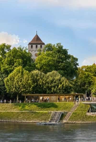 Turm hinter Bäumen am Flussufer mit Menschen auf einem Weg.