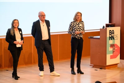 Drei Personen auf einer Bühne bei einer Präsentation mit BAHN 26 Schild.