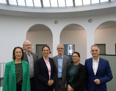 Sechs Personen stehen in einem modernen Innenraum mit Bogenfenstern.