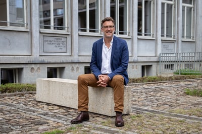 Patrick Langloh sitzt draussen auf Steinbank vor Gebäude mit Fenstern und Inschriften.",