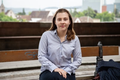 Lilly Simek sitzt auf einer Bank im Freien, Stadt im Hintergrund.