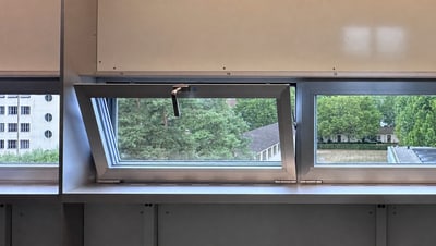 Blick aus einem gekippten Fenster auf Bäume und Gebäude.