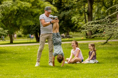 Person hält ein Kind im Handstand im Park, während zwei Kinder zuschauen.