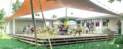 Menschen sitzen auf Terrasse eines Cafés im Freien unter Sonnensegeln.