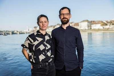Portrait von zwei Personen, links eine Frau, rechts ein Mann. Im Hintergrund sieht man den Rhein und die Skyline von Basel.