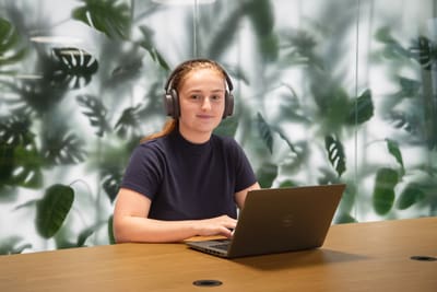 Frau mit Kopfhörern arbeitet am Laptop, Pflanzenhintergrund.",