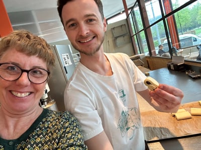 Zwei Personen in einer Bäckerei lächeln mit Teigstücken in der Hand.