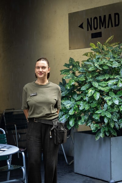 Die Restaurationsfachfrau Manon Furler lächelt im Freien neben Pflanze vor Nomad-Schild.