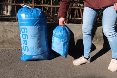 Person hält blauen Abfallsack neben grösserem Sack auf Gehweg.