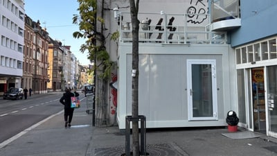 Strassenszene mit Gebäude und Baum, Person trägt Einkaufstüte. Im Zentrum des Bildes sieht man die Luftmessstation Feldbergstrasse.