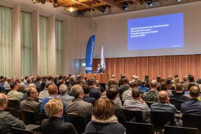 Konferenz mit Publikum und Präsentation auf der Leinwand.",