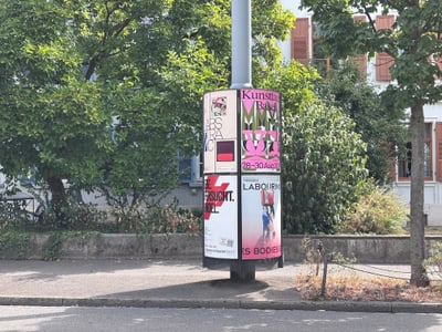 Werbesäule mit Plakaten vor einem Gebäude und Bäumen.