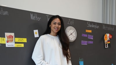 Frau vor Tafel mit Uhr und Beschriftungen in Klassenzimmer.