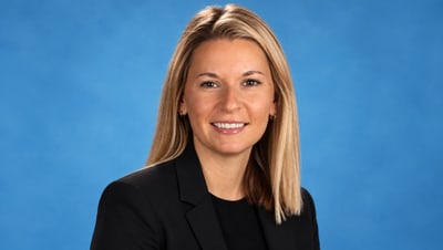 Person mit blonden Haaren in schwarzem Blazer vor blauem Hintergrund.